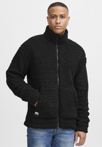 Giacca in pile nera con colletto alto, zip frontale, tessuto texturizzato e due tasche laterali. Patch logo notevole vicino all'orlo.