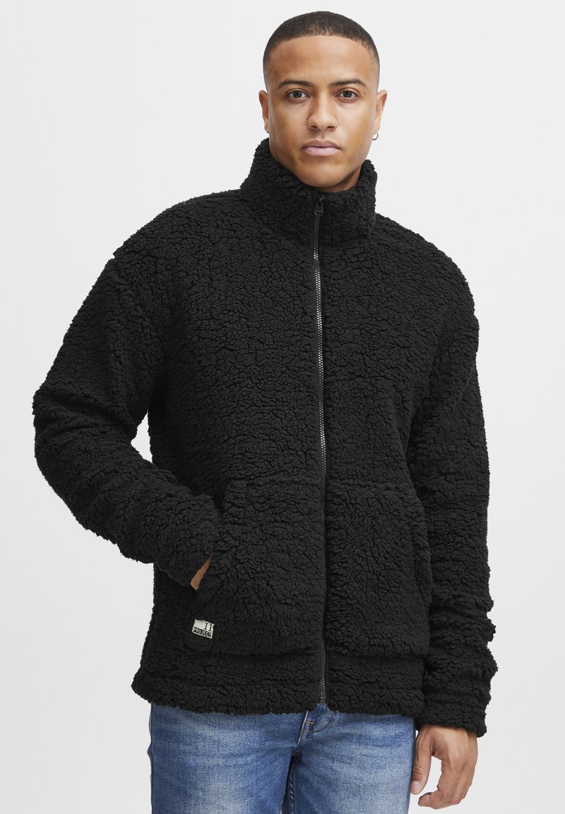Giacca in pile nera con colletto alto, zip frontale, tessuto texturizzato e due tasche laterali. Patch logo notevole vicino all'orlo.