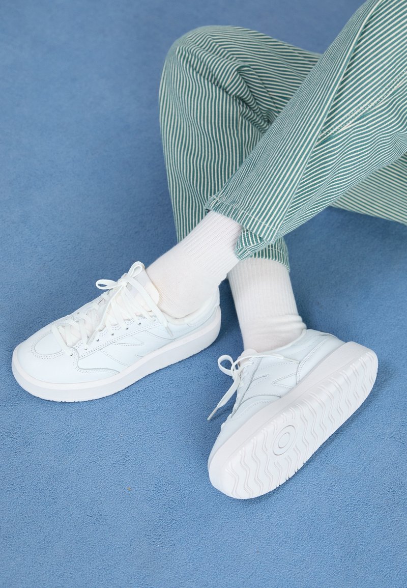 Personne portant des baskets blanches et des chaussettes blanches avec un pantalon à rayures vertes et blanches, assise sur un tapis bleu.