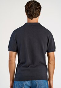 Homme aux cheveux bruns courts portant un polo noir à manches courtes et un jean bleu, vu de dos sur un fond uni.