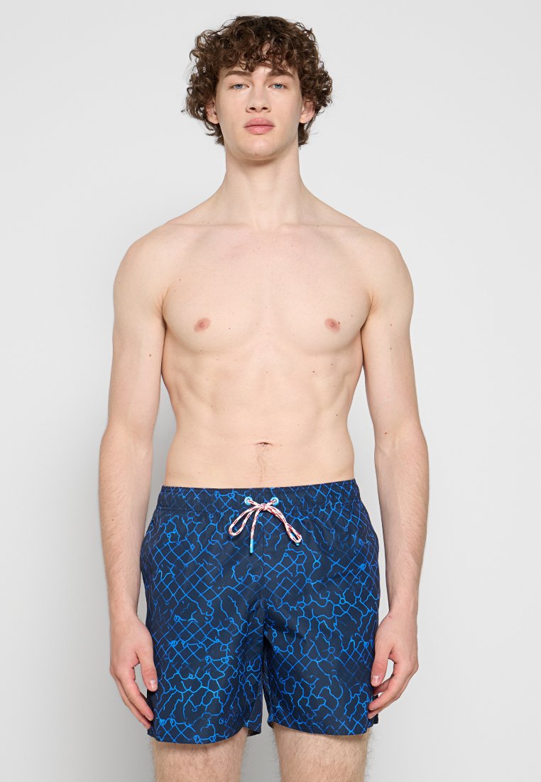 Jeune homme aux cheveux bouclés portant un short de bain bleu à motifs avec un cordon blanc et rouge, debout devant un fond clair uni.