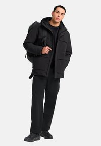 Veste imperméable noire avec une capuche, présentant plusieurs poches avant, portée sur une chemise noire et associée à un pantalon sombre et des chaussures de randonnée.