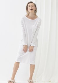 Abito bianco a maniche lunghe con scollo rotondo e tasca sul petto, realizzato in tessuto leggero. Il modello indossa ciabatte beige in peluche.
