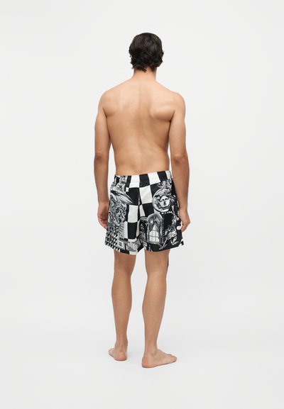 Homme debout, pieds nus, de dos, portant un short à carreaux noir et blanc avec des motifs artistiques de visages et des impressions abstraites.