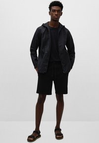 Svart lett hettejakke, svart T-skjorte, svarte shorts og svarte sandaler. Glatt tekstur, minimalistisk design, lommer med klaff.