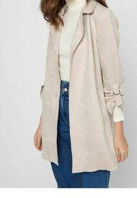 Giacca in suede sintetico di colore beige chiaro con una vestibilità ampia, apertura frontale, e dettagli a fiocco sulle maniche, abbinata a un dolcevita bianco e jeans blu.