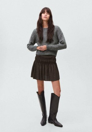 Pull en tricot gris, jupe noire à volants avec taille élastique, et bottes noires hautes au fini brillant. Tenue simple et décontractée.