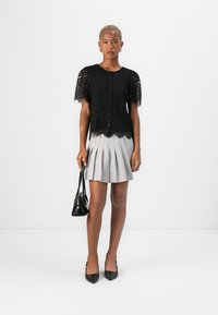 Top corto in pizzo nero con maniche a sbuffo e bottoni sul davanti, abbinato a una gonna grigia plissettata. Modella con una piccola borsa nera e tacchi a punta.