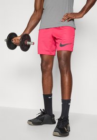 Rózsaszín rövidnadrág fekete Nike logóval, szürke sportfelső, fekete zoknik és fekete edzőcipők, egy kézben súlyzóval.