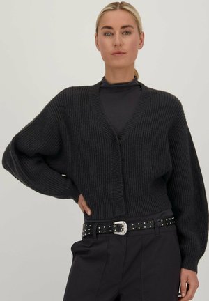 Cardigan tricoté épais gris foncé avec des manches surdimensionnées, décolleté en V profond, fermeture à boutons, porté sur un haut noir et associé à une ceinture cloutée.