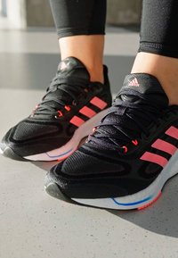 Zwarte sportschoenen met roze accenten, bovenwerk van mesh en een gevoerde zool. Heeft drie roze strepen en oranje veters. Vlakke, gestructureerde zool.