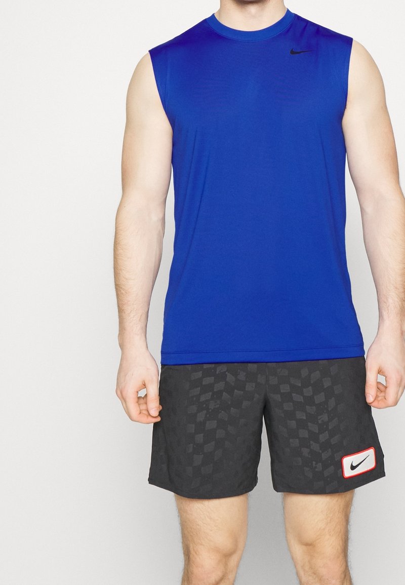 Homme portant un débardeur de sport bleu et un short noir à carreaux avec des logos Nike, debout devant un fond blanc.