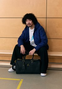 Mala desportiva preto com textura, detalhes em dourado e um logótipo. Modelo veste um casaco acolchoado azul-marinho, t-shirt branca com estampa gráfica e calças pretas.