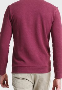 Man i långärmad vinröd tröja och beige byxor sedd bakifrån, stående mot en enfärgad bakgrund.