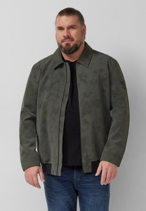 Bärtiger Mann mit zurückgebundenem Haar, der eine dunkelgrüne Jacke mit Reißverschluss über einem schwarzen Hemd und blauen Jeans vor einem schlichten grauen Hintergrund trägt.