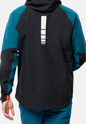 Personne portant une veste noire et bleu sarcelle avec un motif géométrique blanc vertical dans le dos et un pantalon bleu, vue de dos.