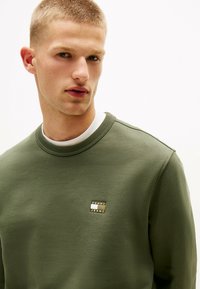 Felpa verde khaki realizzata in tessuto strutturato. Presenta un colletto a girocollo e una piccola etichetta del marchio sul petto. Design minimalista.