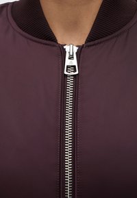 Personne portant une veste bordeaux foncée avec une fermeture éclair argentée proéminente et un col côtelé, montrée du cou jusqu'à la partie supérieure de la poitrine.
