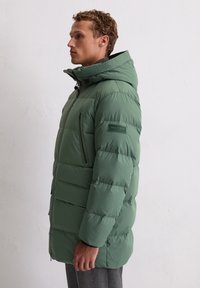 Jasnopistacjowa kurtka puchowa z kapturem, zapinana na zamek błyskawiczny z przodu i posiadająca boczne kieszenie, wyróżniająca się teksturowanym kołnierzem i naszywką z logo na lewym ramieniu.