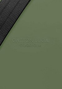 Material verde cu o finisare texturată și o curea neagră, având un branding embossat "GASTON LUGA STOCKHOLM." Design minimalistic.