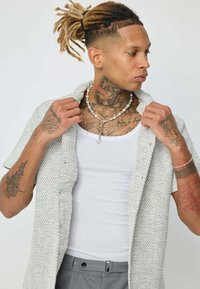 Chemise à manches courtes, de couleur gris clair, avec un tissu texturé ; portée par-dessus un débardeur blanc, mettant en valeur des tatouages complexes sur les bras et le cou.