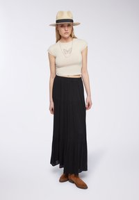 Gonna maxi nera con balze a strati, abbinata a una t-shirt corta color crema con design di farfalla, completata da un cappello di paglia e stivali marroni.