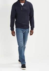 Man som går iklädd en mörk marinblå sjalkrage tröja, ljusblå jeans och svarta sneakers med vita sulor mot en enfärgad vit bakgrund.