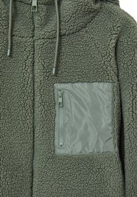 Giacca in fleece verde tenue, con un esterno texturizzato e morbido, una tasca in tessuto liscio con cerniera e coulisse regolabili sul cappuccio.