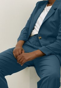 Komplet w kolorze morskim, składający się z marynarki i spodni, wykonany z gładkiego materiału. Marynarka posiada jeden złoty guzik. Nosi się ją na białą koszulkę, ręce spoczywają na kolanach.