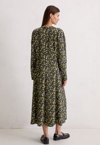 Langsleeve, zwarte jurk met een gele abstracte print, vloeiende silhouet, plissé en ronde halslijn. G gedragen met zwarte platte schoenen.