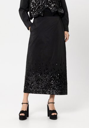 Lange schwarze Rock mit einem mit Pailletten verzierten Saum, der eine glatte Stofftextur aufweist. Kombiniert mit schwarzen Sandalen mit Absatz und Knoten-Detail.