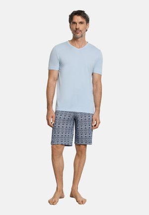 Mann steht barfuß und trägt ein hellblaues Kurzarmhemd sowie gemusterte marineblaue knielange Shorts vor weißem Hintergrund.