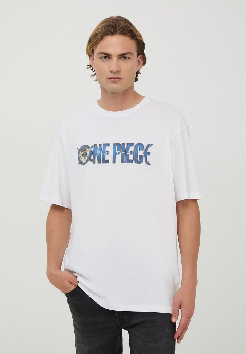 T-shirt bianco in cotone con una grafica "One Piece" in blu e grigio, dal taglio rilassato e maniche corte. Il modello sta in piedi con le mani nelle tasche.