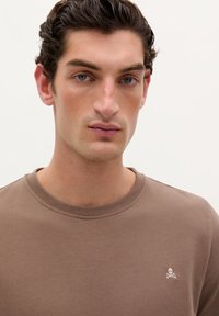 Bruine sweatshirt met een ronde hals, gemaakt van een gestructureerde stof. Bevat een klein wit doodskop- en gekruiste botten logo op de linkerborst.