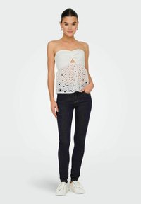 Top bianco senza spalline con un nodo al centro, caratterizzato da un design a fori simile a pizzo. Abbinato a jeans skinny scuri e sneakers bianche.
