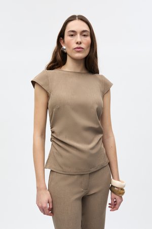 Vrouw draagt taupekleurige top met kapmouwtjes en bijpassende broek, met grote zilveren oorbel en chunky armbanden aan rechterpols, tegen een effen achtergrond.