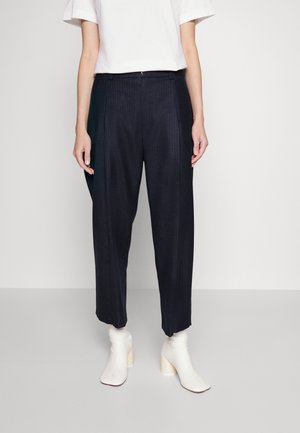 Personne portant un pantalon large court à fines rayures noires, un haut blanc à manches courtes et des bottines blanches à talons, debout sur un sol clair.