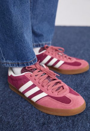 Nahaufnahme von Füßen, die pinke Adidas-Sneaker mit weißen Streifen und Gummisohlen tragen, kombiniert mit blauen Jeans auf einem blauen Teppich.