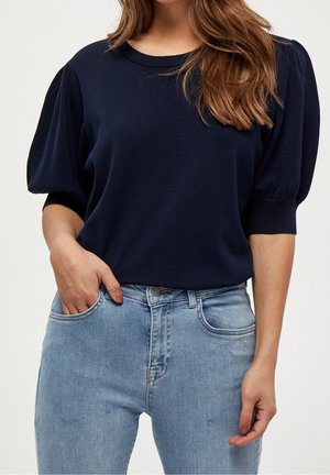 Vrouw draagt een marineblauwe trui met pofmouwen en lichtblauwe jeans, met één hand in haar zak en los golvend haar over haar schouder.