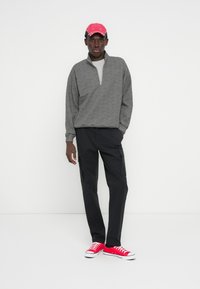 Sweat-shirt gris à fermeture éclair, couche intérieure grise, pantalon cargo noir et baskets rouges. La casquette est rose vif avec des détails blancs.