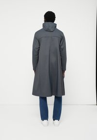 Halli veekindel jakk kapuutsiga, millel on pikk kuju, minimaalne disain ja sile tekstuur, kantud siniste teksade ja valgete tennisetega.