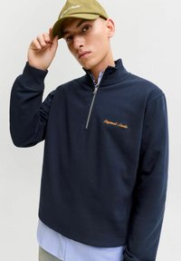 Tumedateed neljakannaga fliis, ribakäistega; sisaldab kuldset tikanditeksti. Kantud helesinise kaelusega särgi peale. Vaba lõige.