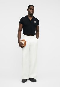 Homme portant un polo noir raccourci, un pantalon large blanc, des chaussures noires, tenant une petite pochette marron avec une main dans la poche.