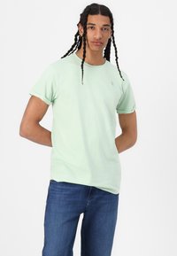 Jasnozielona bawełniana koszulka z krótkim rękawem i podwiniętymi mankietami, z małym logo na klatce piersiowej. Noszona z niebieskimi dżinsami.