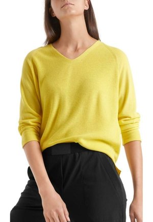 Femme portant un pull jaune vif à col en V avec manches retroussées et un pantalon noir taille haute sur fond blanc.