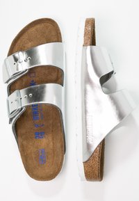 Sølvmetallic sandaler med to justerbare spænder, en brun ruskinds fodseng og en hvid gummisål, som har en tekstureret overflade.