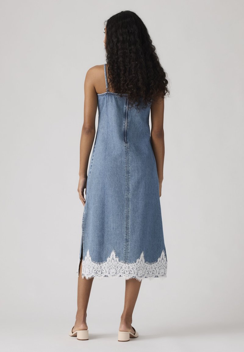 Frau mit langen lockigen Haaren, die ein blaues Denim-Midikleid mit weißem Spitzenbesatz am Saum trägt, steht mit dem Rücken zum Betrachter vor einem einfarbigen Hintergrund.