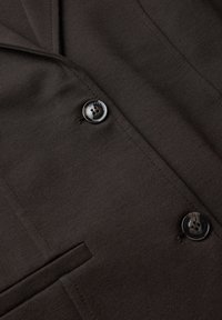 Gros plan sur un blazer marron foncé montrant deux boutons noirs, un revers, un tissu texturé et une partie d’une poche passepoilée.