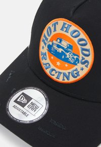 Casquette noire avec un patch rond orange et beige portant le texte "HOT HOODS RACING" et un design de camion vintage bleu. Détail de l'érable réglable visible.