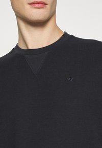 Gros plan d'un homme portant un pull noir à col rond côtelé avec un petit logo brodé représentant des pagaies croisées sur la poitrine.